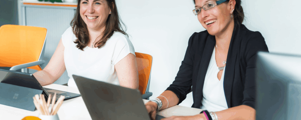 Zwei Frauen sitzen mit Laptops an einem Tisch im Buero