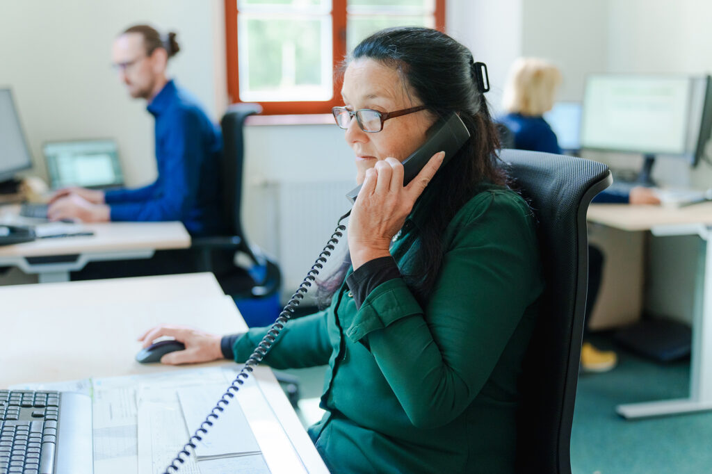 Eine Mitarbeiterin der Benke Steuerberatung telefoniert an ihrem Arbeitsplatz, während Kollegen im Hintergrund am Computer arbeiten.