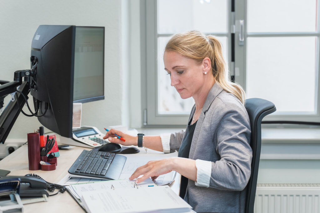 Eine Mitarbeiterin der Benke Steuerberatung arbeitet konzentriert am Schreibtisch, sortiert Unterlagen und nutzt Computer und Taschenrechner.