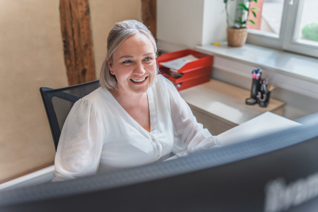 Eine lächelnde Mitarbeiterin der Benke Steuerberatung sitzt an ihrem Schreibtisch und arbeitet am Computer in einem hellen Büro.