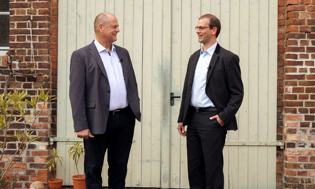 Der Gründer und der Nachfolger der Benke Steuerberatung stehen gemeinsam vor einer Scheunentür, unterhalten sich und lächeln einander zu.