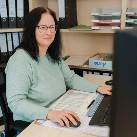 Eine Mitarbeiterin der Benke Steuerberatung arbeitet am Computer, nutzt Unterlagen und lächelt leicht, während sie an ihrem Schreibtisch sitzt.