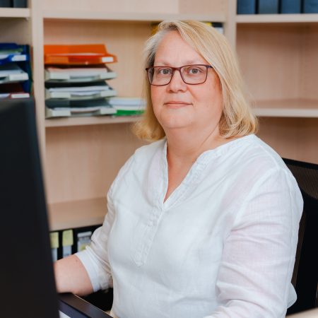 Eine Mitarbeiterin der Benke Steuerberatung sitzt an ihrem Schreibtisch und blickt freundlich in die Kamera, im Hintergrund sind Aktenregale zu sehen.