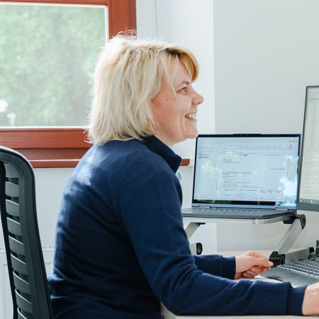 Eine Mitarbeiterin der Benke Steuerberatung sitzt an ihrem Schreibtisch, arbeitet am Laptop und lächelt dabei.