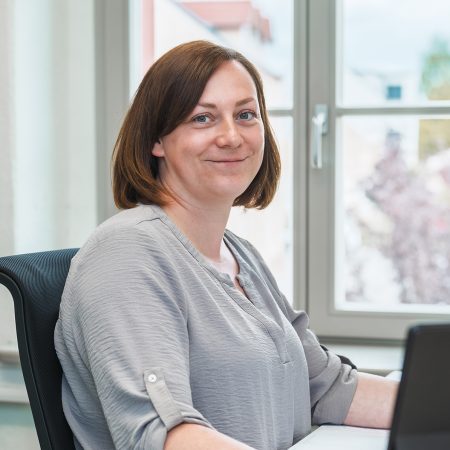 Eine Mitarbeiterin der Benke Steuerberatung sitzt an ihrem Arbeitsplatz vor dem Laptop und lächelt freundlich in die Kamera.